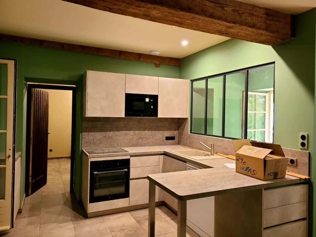 Cuisine toute équipé, murs verts, charpente en bois, verrière au mur, et luminaires allumés.